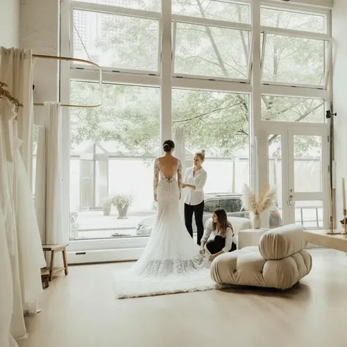 Una sposa sorridente mentre prova uno splendido abito da sposa in una boutique di lusso, assistita da due consulenti nuziali professioniste.