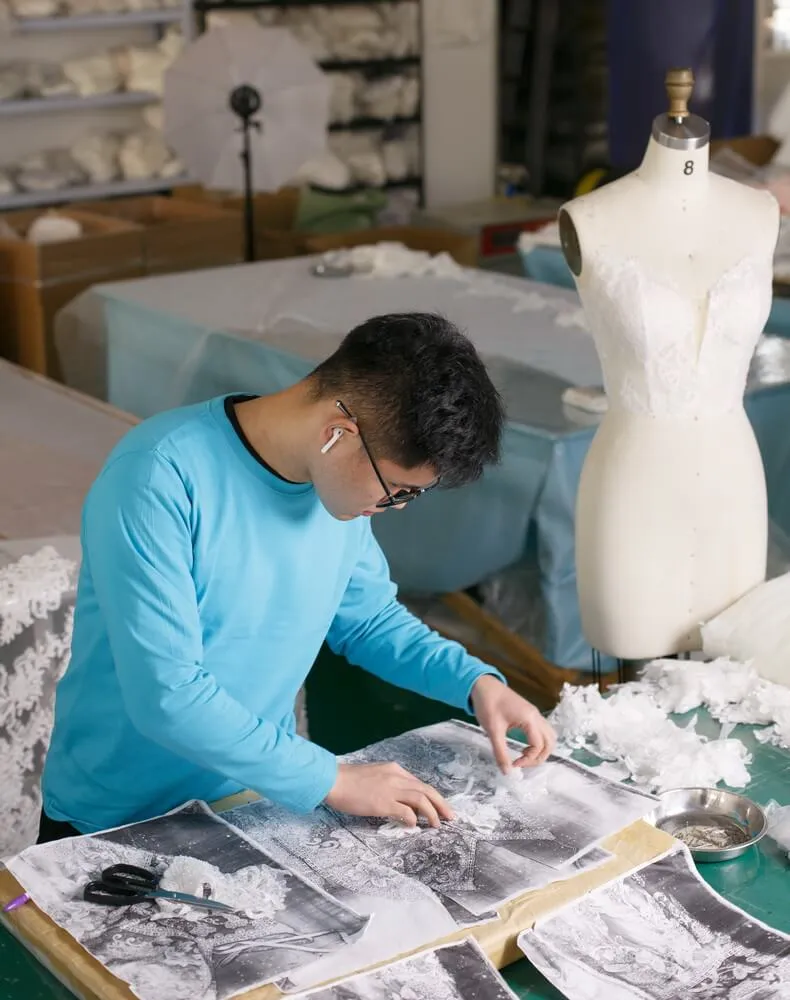 A designer meticulously crafting lace components for a wedding dress at a workbench with design sketches, showcasing the process from concept to handcrafted reality.