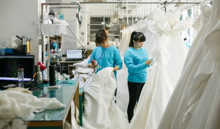 Deux artisans qualifiés confectionnent des robes de mariée haute couture dans un atelier de robes de mariée lumineux et bien organisé, avec des rangées de robes finies en arrière-plan illustrant le processus de fabrication professionnel.